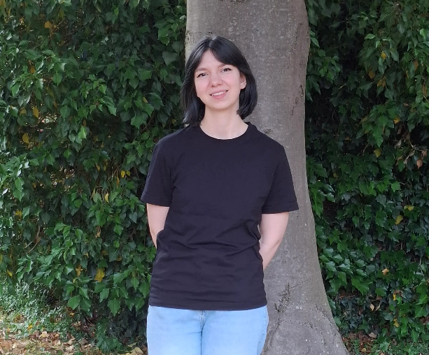 A person with short dark hair stands in front of a tree, wearing a black T-shirt and light blue jeans, with dense greenery in the background.