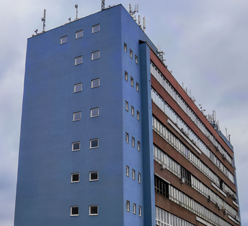 A tall building with a blue facade and many antennas on the roof, featuring rows of square and rectangular windows against a cloudy sky.