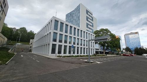 Modern white building with large windows on a street corner, surrounded by taller glass buildings and trees under a cloudy sky.