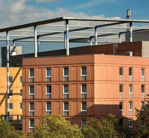 A modern building with orange and yellow sections, featuring a metal framework on the roof, surrounded by trees under a partly cloudy sky.