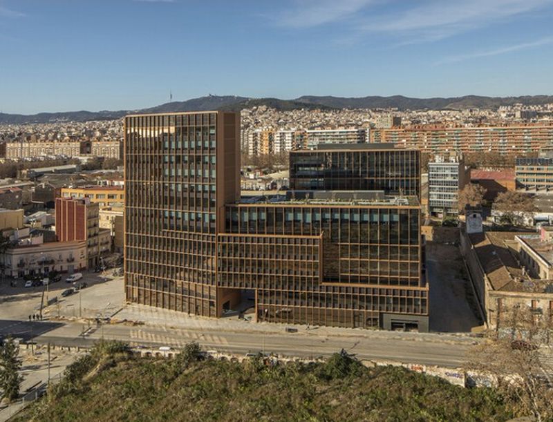 Aerial view of a modern, multi-story office building with glass windows and brown exterior, set against a cityscape with distant hills.