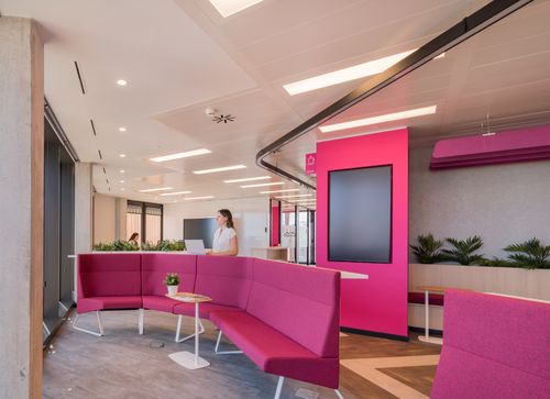 Modern office lounge with vibrant pink seating, indoor plants, a digital screen, and a person working on a laptop at a desk.
