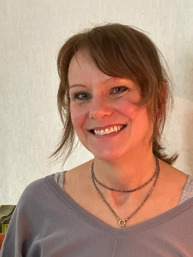 Smiling woman with brown hair, wearing a gray top and layered necklaces, against a light background.