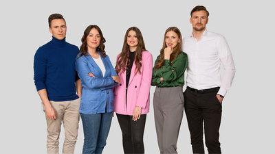 Five people standing in a row against a gray background, wearing casual and semi-formal attire, smiling at the camera.