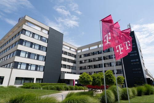 Modern office building with black and white facade, surrounded by greenery. Red flags with a white "T" logo are flying in the foreground.