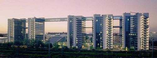 Modern office complex with multiple connected high-rise buildings, featuring glass facades and a sky bridge, set against a twilight sky.