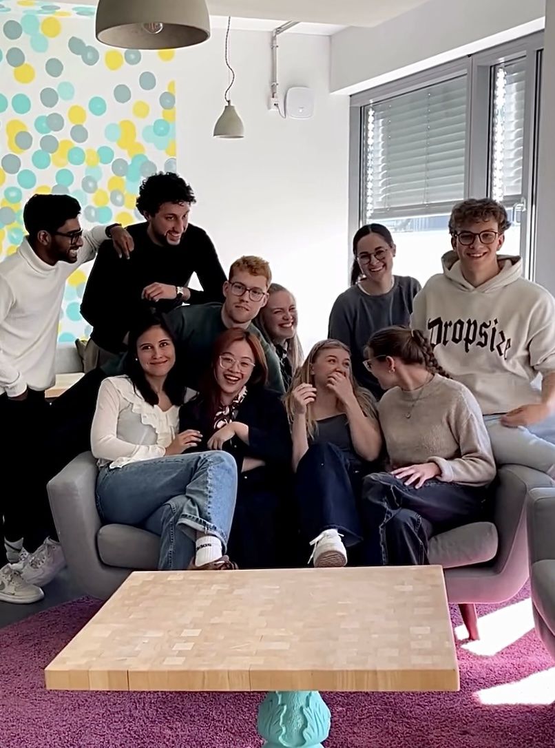 A group of young people smiling and sitting together on a sofa in a modern room with colorful polka dot wallpaper and large windows.
