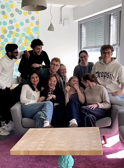 A group of young people smiling and sitting together on a sofa in a modern room with colorful polka dot wallpaper and large windows.