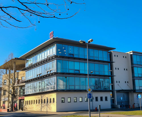 Modern glass office building with a red logo on the roof, set against a clear blue sky, surrounded by bare trees and street signs.