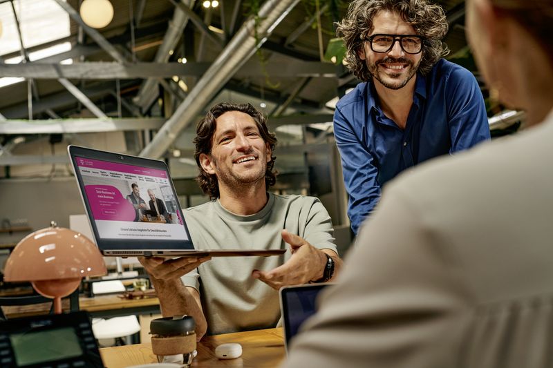 Two men smiling and showing a laptop screen to a woman in an office setting, with modern decor and natural lighting.