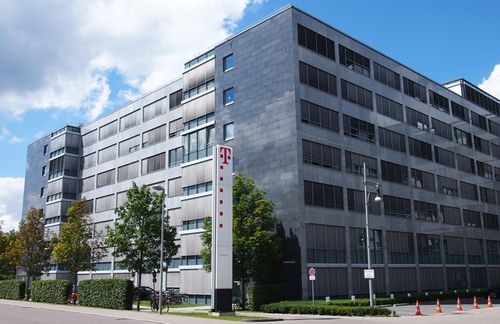 Modern office building with a Telekom logo, surrounded by trees and a clear sky. Orange cones line the sidewalk.