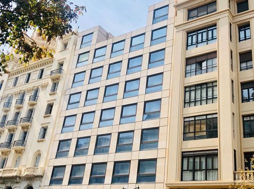 Modern beige building with large windows, flanked by two older, ornate buildings, under a clear blue sky with tree leaves in the foreground.