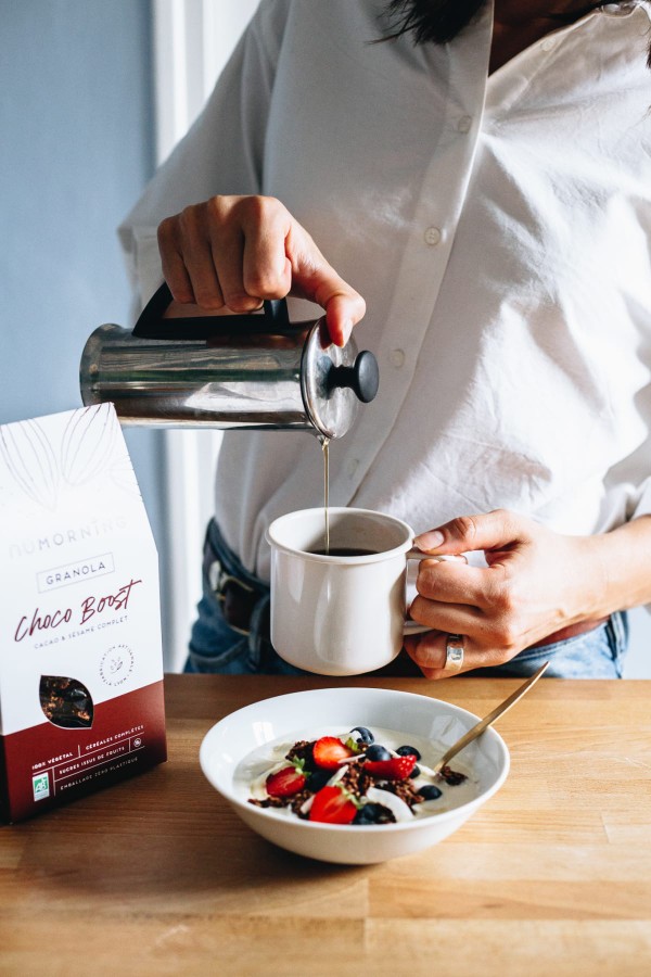 Jeune femme se servant du café, devant une table avec un bol de granola aux fruits frais