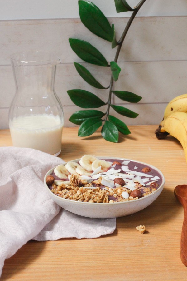 Açaï bowl avec toppings de banane et copeaux de noix de coco, servi sur une table avec une grappe de bananes et un carafe de lait végétal