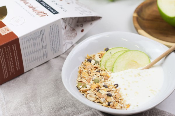 Bol de muesli avec tranches de pommes, yaourt végétal, pépites de chocolat et graines de courge à côté d'un paquet de muesli au chocolat