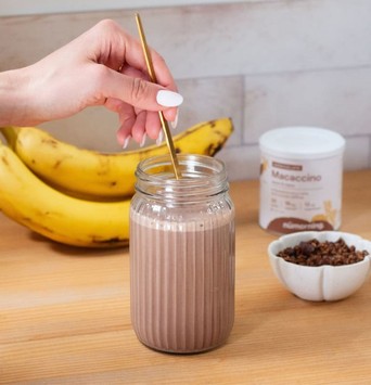 Smoothie végan au chocolat dans un grand verre, remué par une main de femme, avec une grappe de bananes et un pot de chocolat en poudre en arrière-plan.