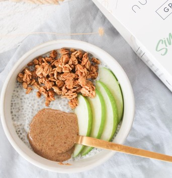 Bol de pudding de chia avec granola, cuillère de beurre de cacahuète et tranches de pomme, sur une table de petit-déjeuner avec un paquet de granola