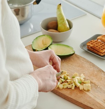 Jeune femme découpant un avocat en petits morceaux sur une planche à découper, à côté d'une assiette de fruits et d'une autre de gauffres