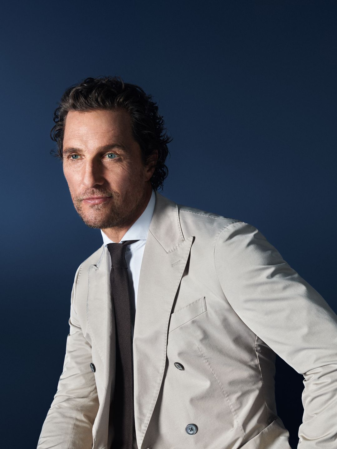 Matthew McConaughey trägt ein weißes OLYMP-Hemd mit beigen Anzug und dunkler Krawatte vor dunklem Hintergrund.