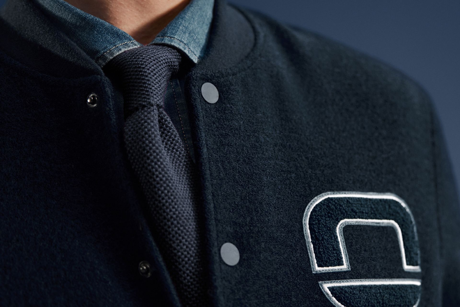 Nahaufnahme eines Mannes in dunklblauer Collegejacke mit gesticktem OLYMP-Logo, darunter Hemd und blauer Krawatte.