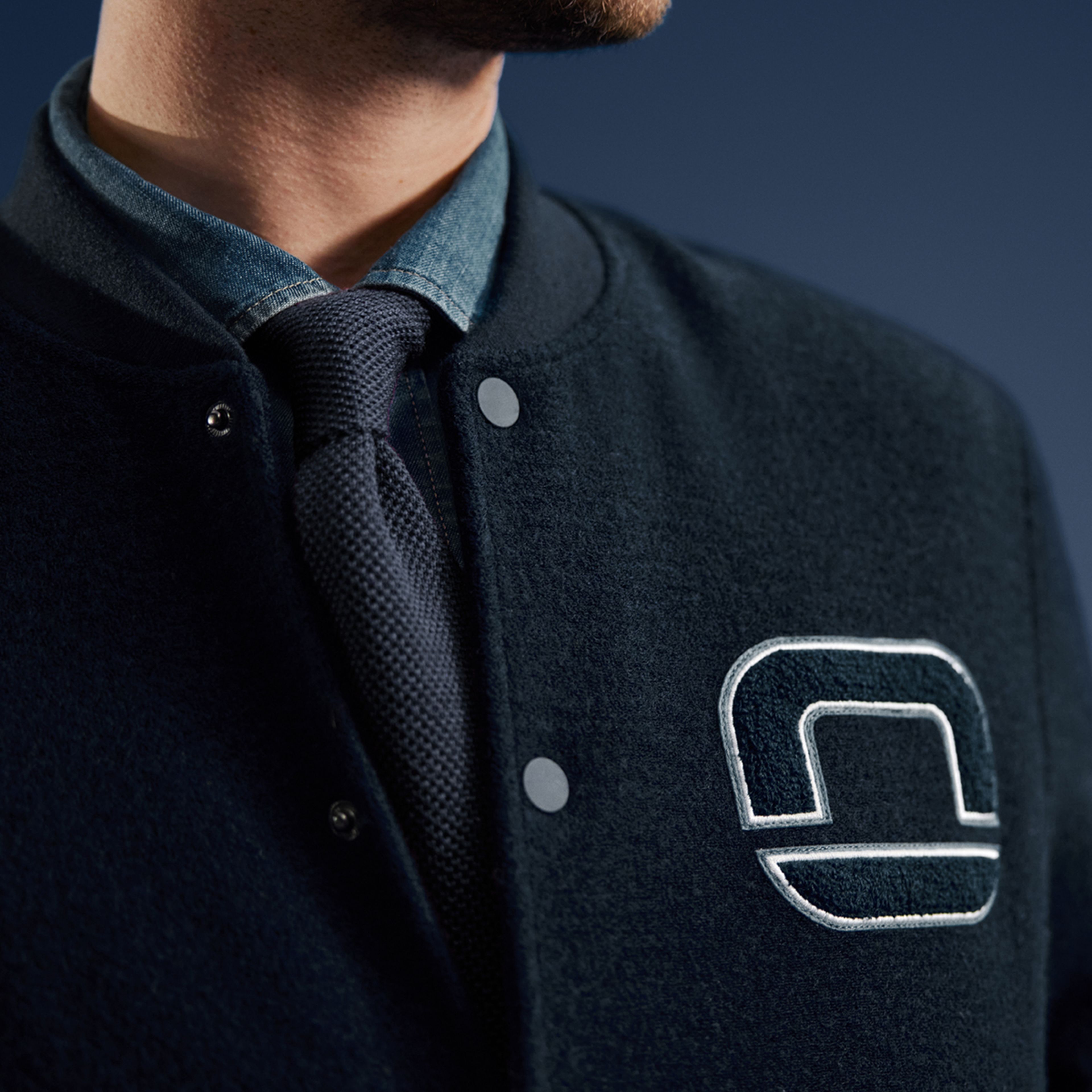 Nahaufnahme eines Mannes im dunklblauer Collegejacke mit gesticktem OLYMP-Logo, darunter Hemd und blaue Krawatte