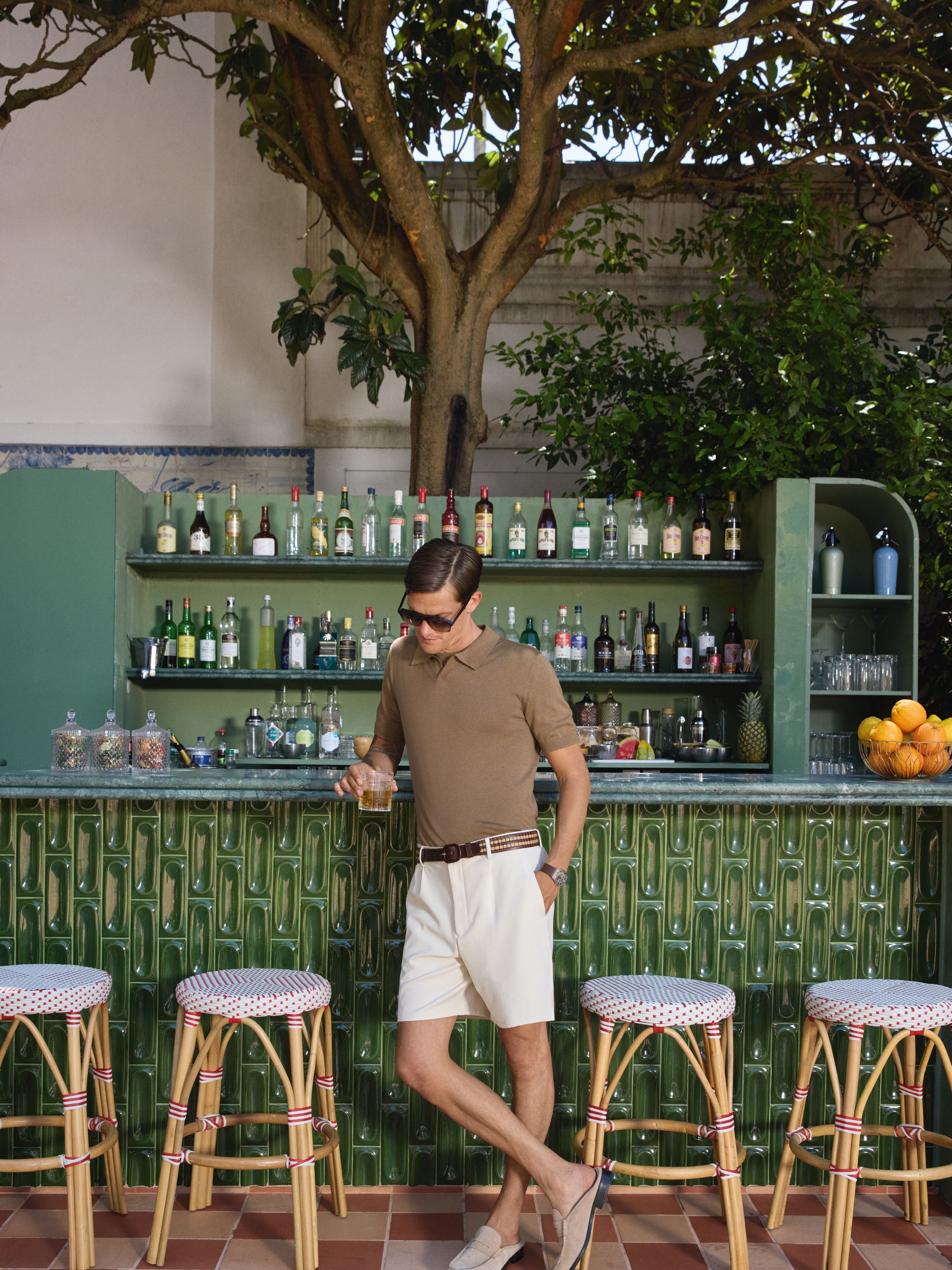 Männliches Model steht lässig an einer grünen Outdoor-Bar unter einem Baum. Er trägt ein sandfarbenes OLYMP-Poloshirt, helle Shorts und hält ein Getränk in der Hand.