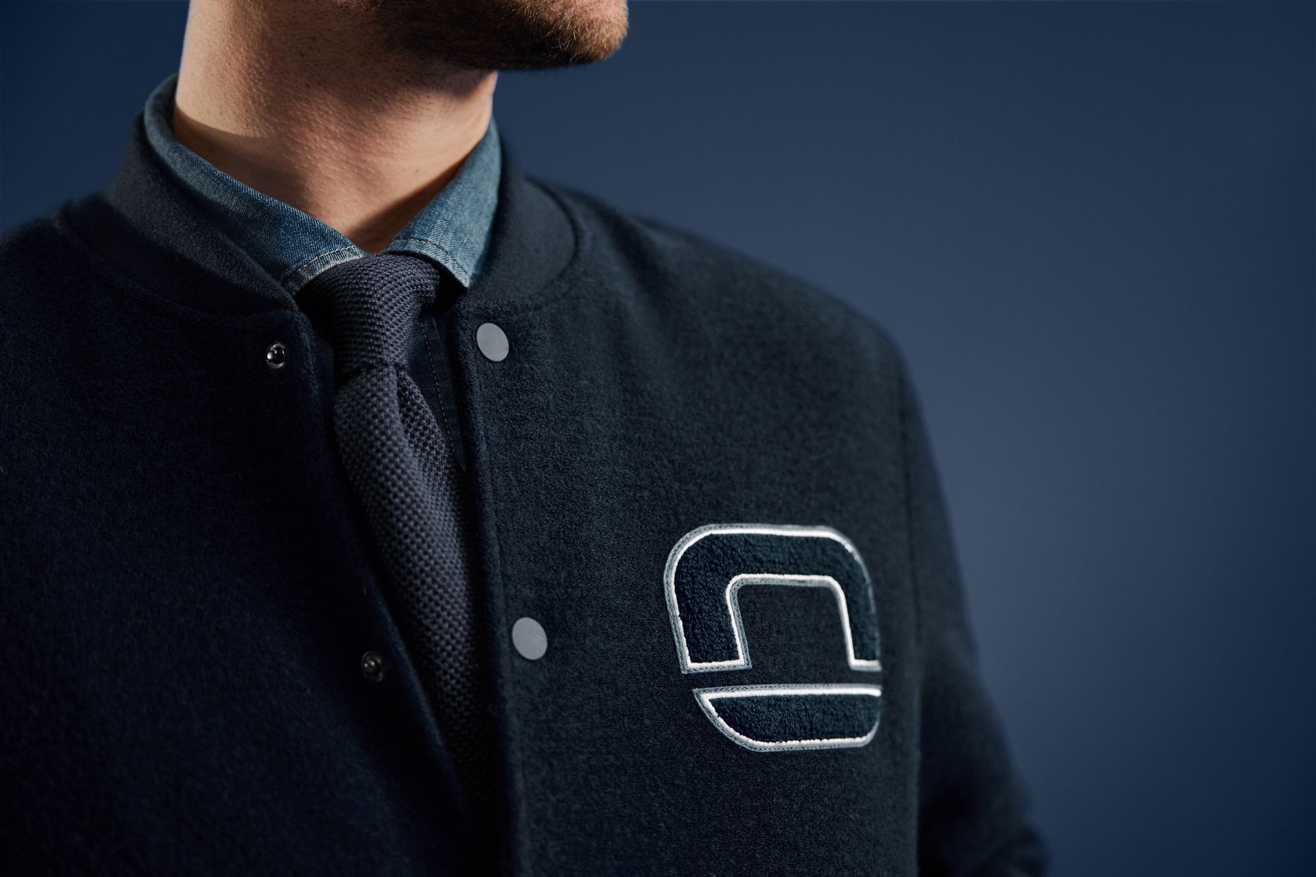Nahaufnahme eines Mannes in dunklblauer Collegejacke mit gesticktem OLYMP-Logo, darunter Hemd und blaue Krawatte