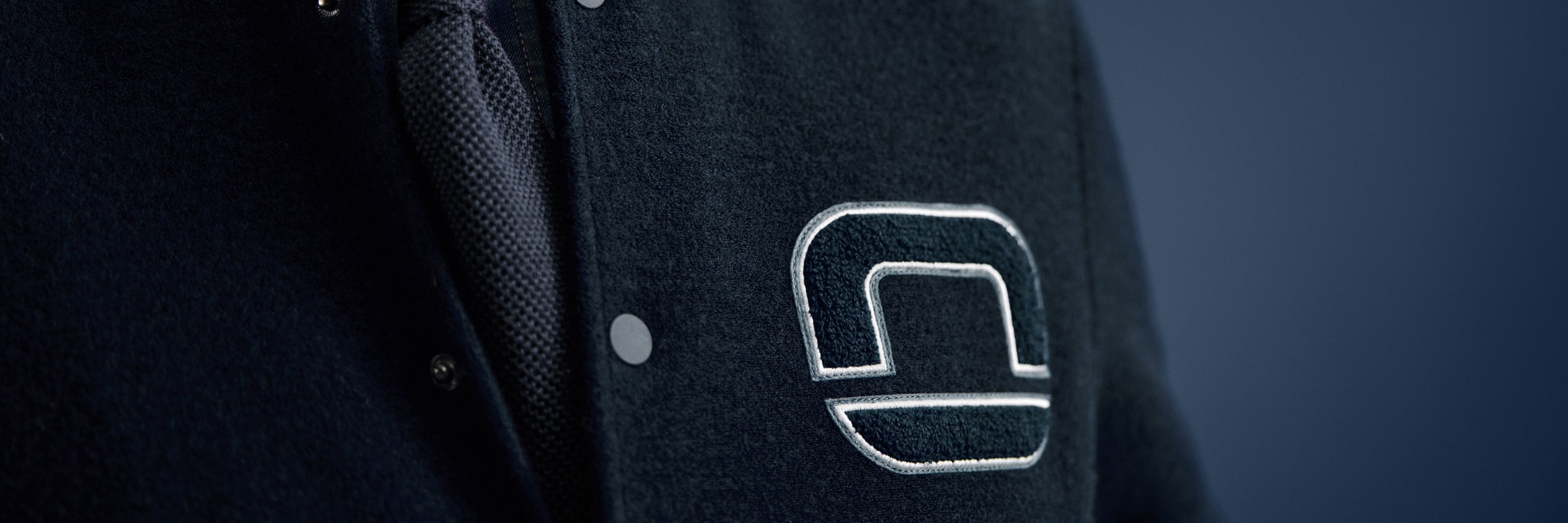 Nahaufnahme eines Mannes in dunklblauer Collegejacke mit gesticktem OLYMP-Logo, darunter Hemd und blauer Krawatte.
