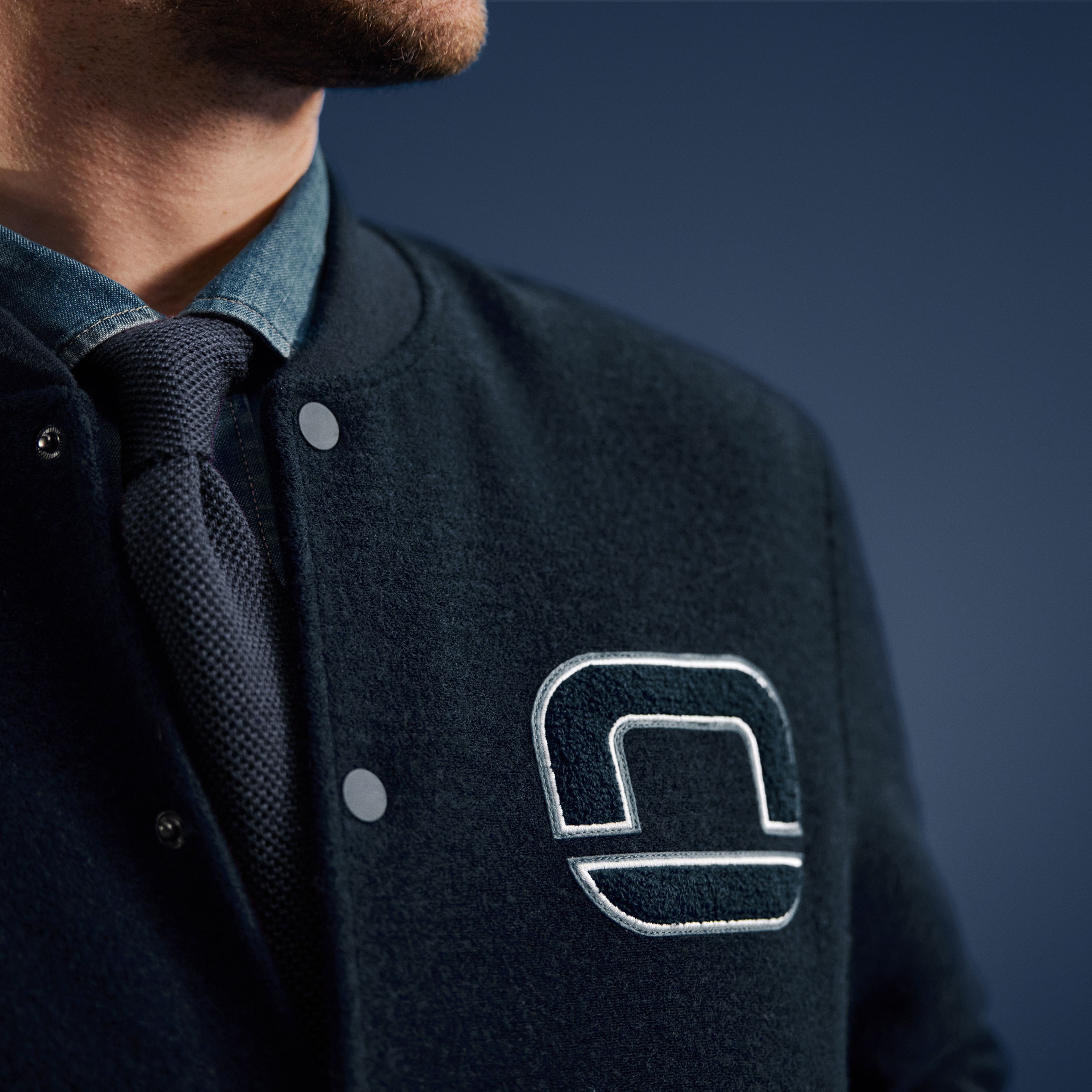 Nahaufnahme eines Mannes in dunklblauer Collegejacke mit gesticktem OLYMP-Logo, darunter Hemd und blauer Krawatte