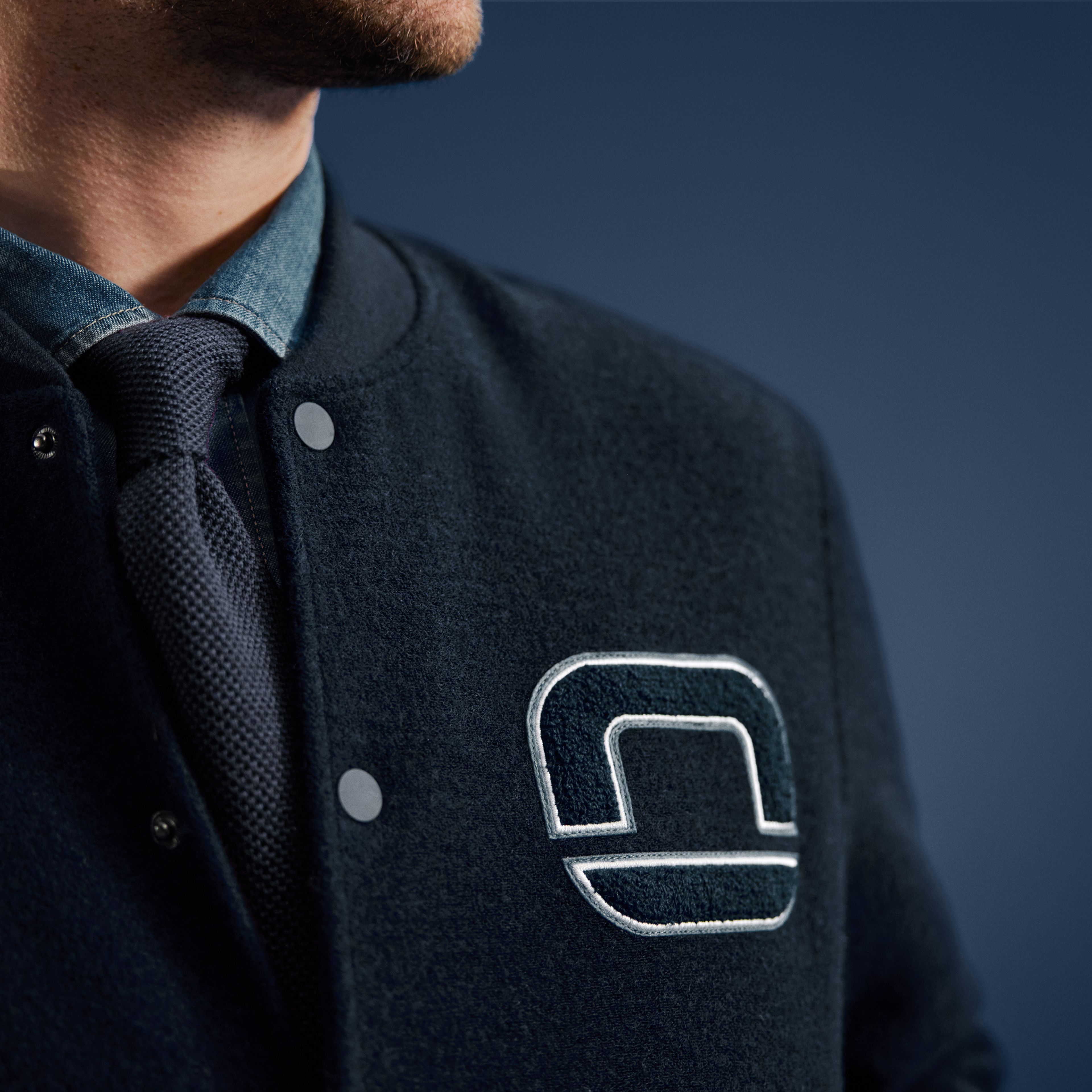 Nahaufnahme eines Mannes in dunklblauer Collegejacke mit gesticktem OLYMP-Logo, darunter Hemd und blaue Krawatte