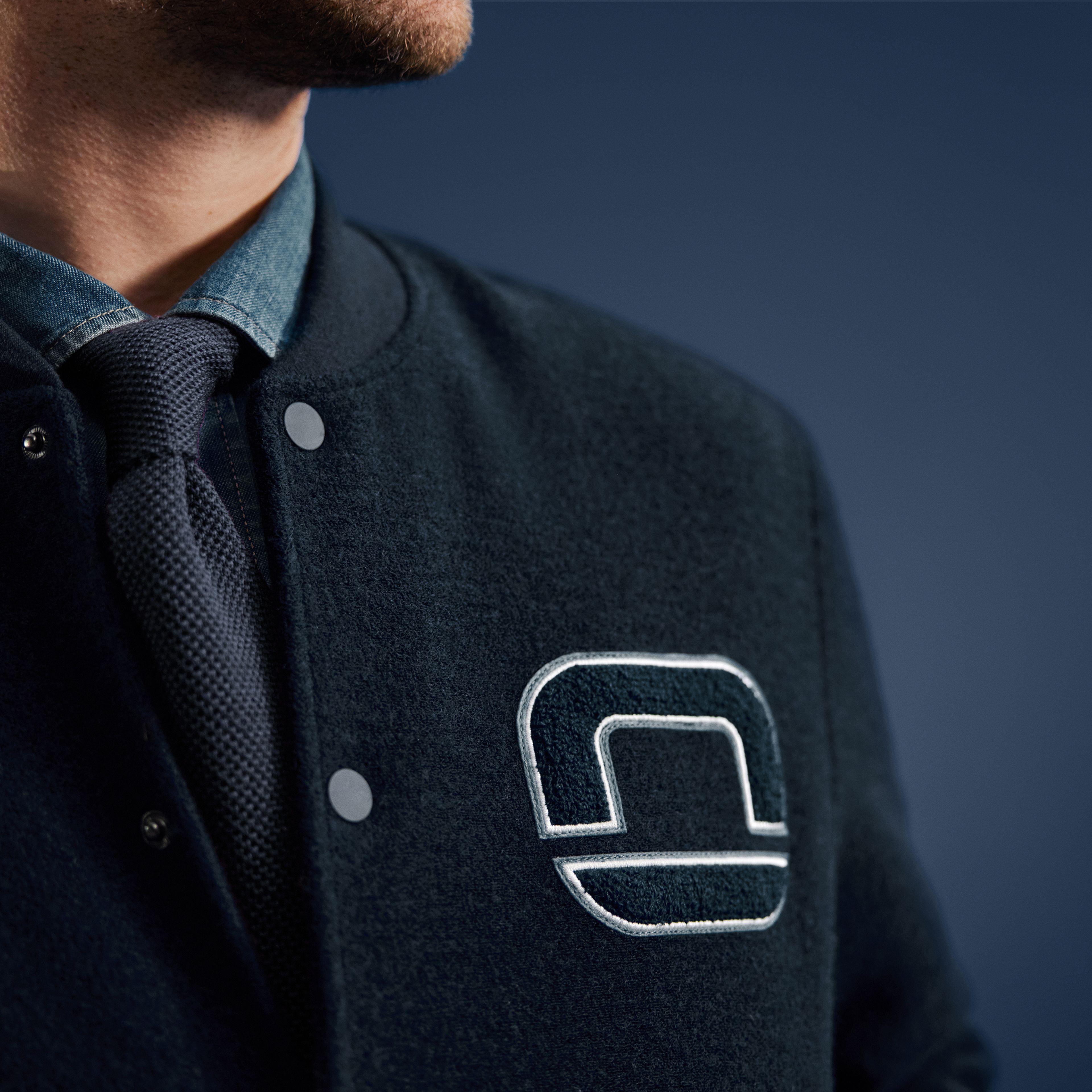 Nahaufnahme eines Mannes in dunklblauer Collegejacke mit gesticktem OLYMP-Logo, darunter Hemd und blaue Krawatte