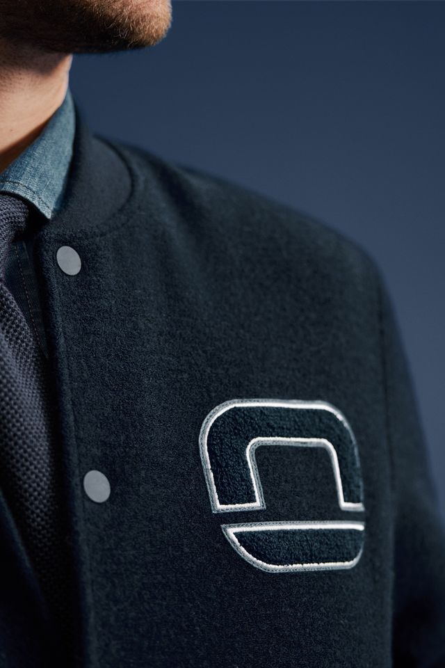 Nahaufnahme eines Mannes in dunklblauer Collegejacke mit gesticktem OLYMP-Logo, darunter Hemd und blaue Krawatte