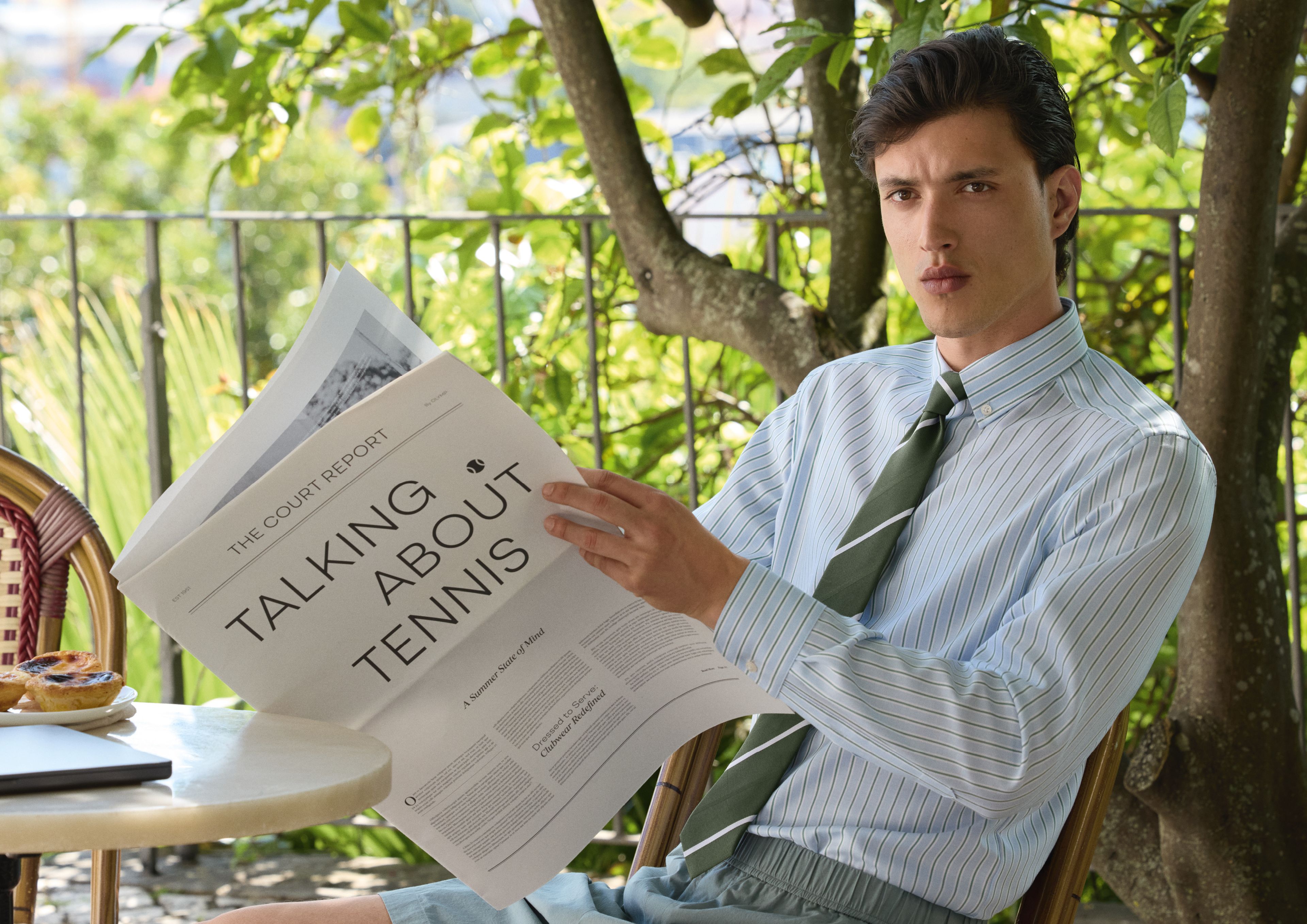 Männliches Model der OLYMP Frühjahr Sommer 2026 Kollektion sitzt in einem Café und liest eine Zeitung. Er trägt ein hellblau gestreiftes Hemd mit grüner Krawatte.