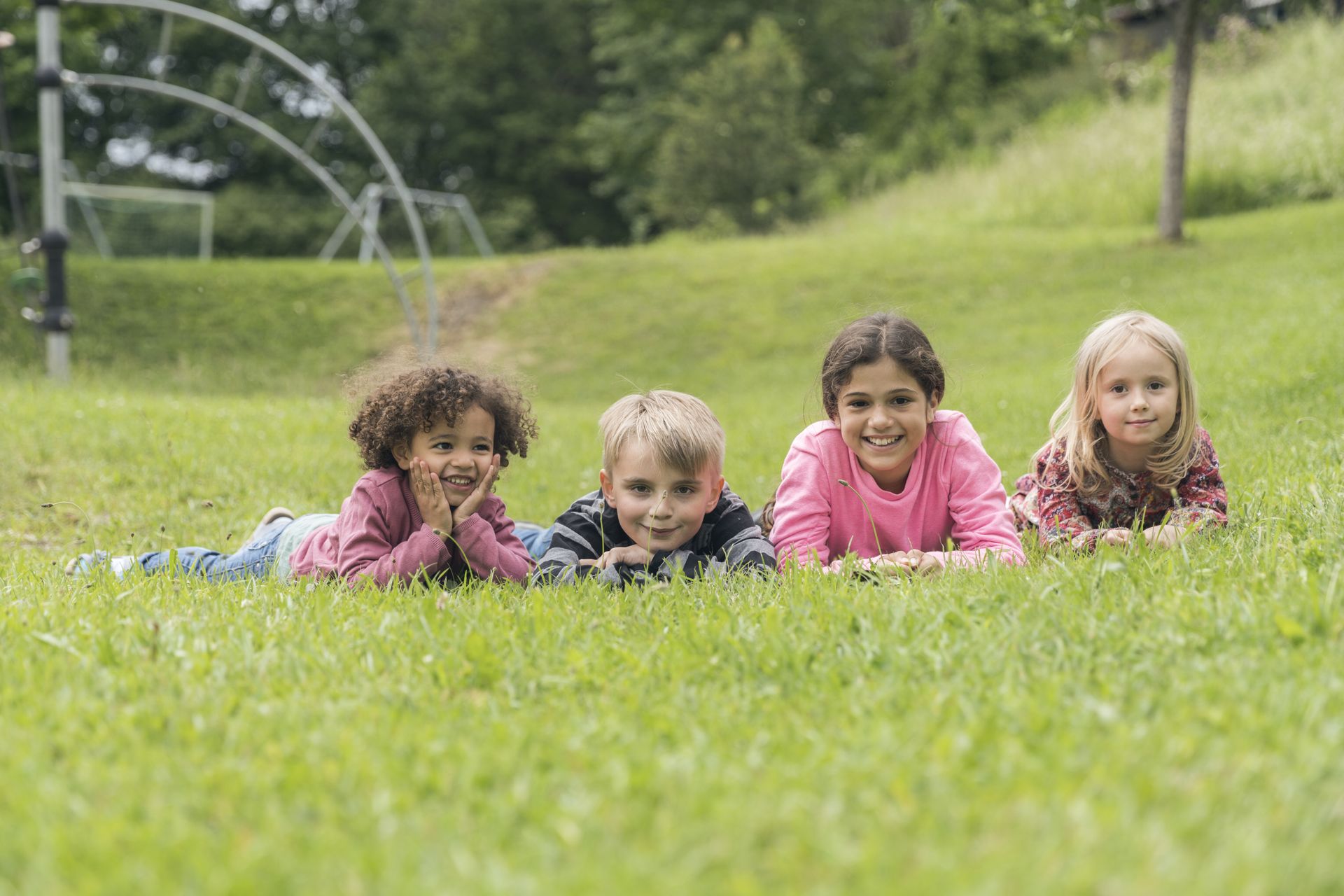 Vier Kinder liegen lachend nebeneinander auf einer grünen Wiese.