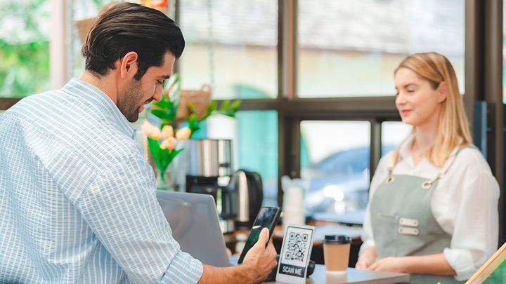 Ein Mann scannt an der Theke eines Cafés mit seinem Handy einen QR-Code, während eine Frau in einer Schürze hinter der Theke steht und zusieht.