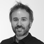Black and white professional portrait of Julien Mangas, Interim CTO at LINK Mobility Group. He is smiling, has medium-length tousled hair and a beard, and is wearing a dark collared shirt. The background is plain and softly lit.