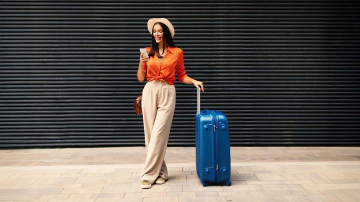 Eine Frau in einem orangefarbenen Shirt und einer beigen Hose, die einen Hut trägt, an einem blauen Koffer lehnt, lächelt und auf ihr Handy schaut, vor dem Hintergrund eines schwarzen Rollladens.
