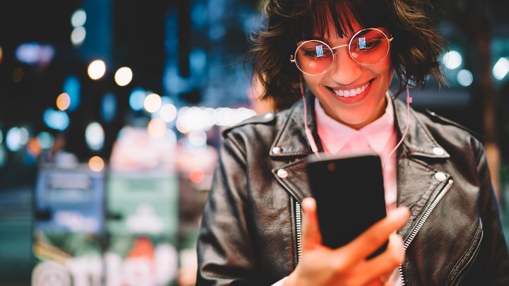Eine Frau in einer Lederjacke, die lächelnd auf ihr Handy schaut, eine Brille und Kopfhörer trägt, vor dem Hintergrund einer verschwommenen nächtlichen Stadtansicht.