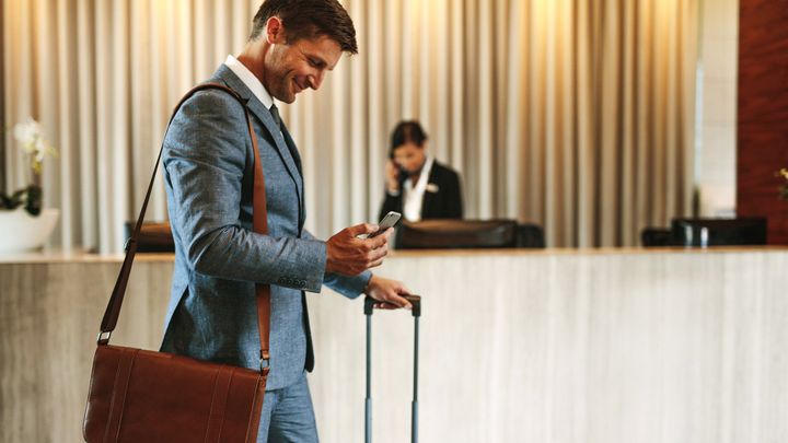 Ein Mann im Anzug, der auf sein Handy schaut, Gepäck in der Hand hält und eine Ledertasche über der Schulter trägt, steht in einer Hotellobby. Im Hintergrund ist eine Rezeptionistin zu sehen.