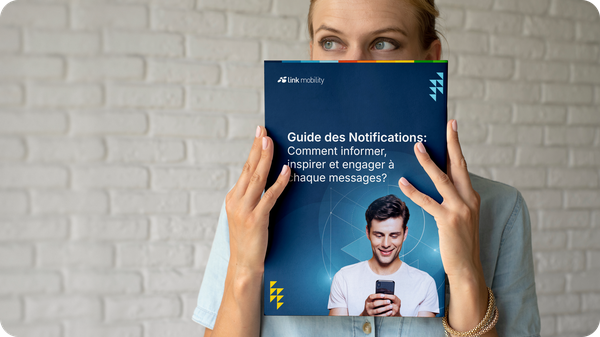 Une personne tient devant un mur de briques blanches une brochure intitulée « Guide des Notifications » de « Link Mobility », qui lui cache partiellement le visage.
