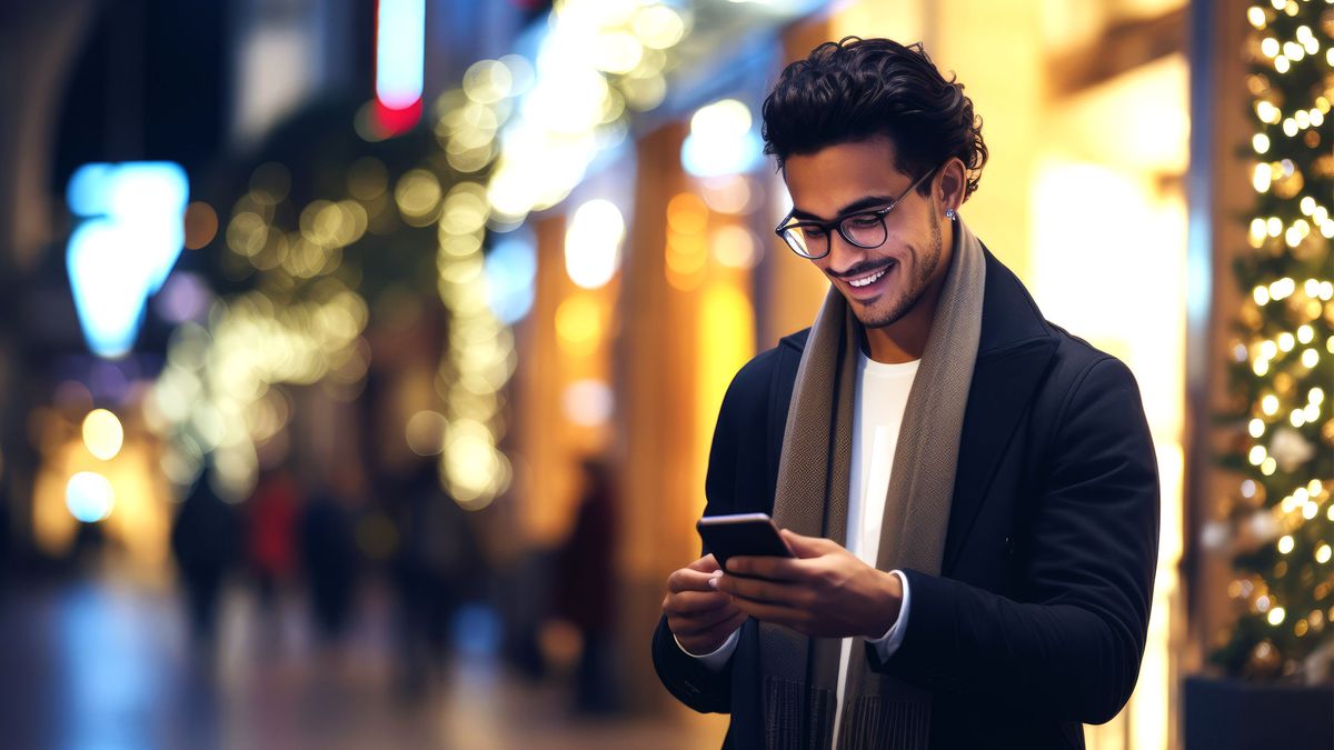 Ein Mann mit Brille und Schal lächelt, während er nachts auf einer hell erleuchteten Straße sein Smartphone benutzt; im Hintergrund sind verschwommene Lichterketten zu sehen.