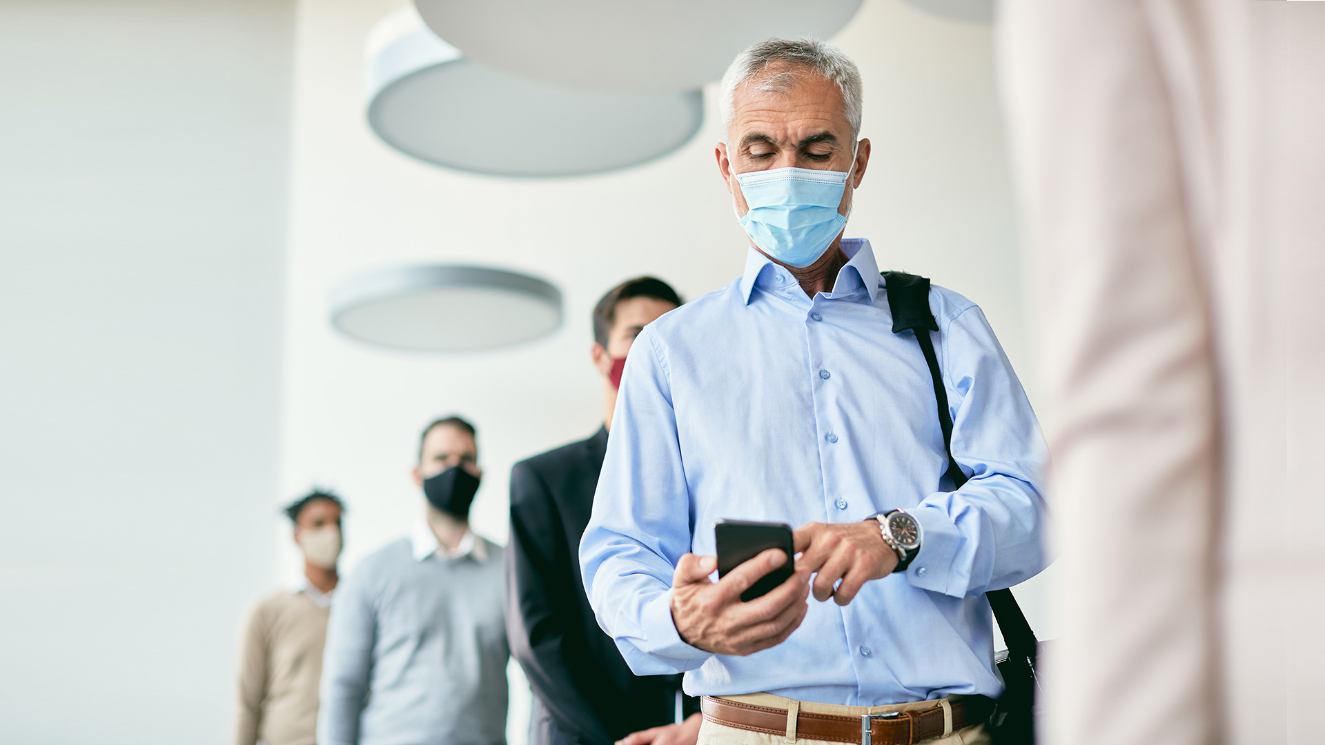 Eine Gruppe von Menschen mit Masken steht in einem Raum in einer Reihe. Der Mann im Vordergrund benutzt ein Smartphone.