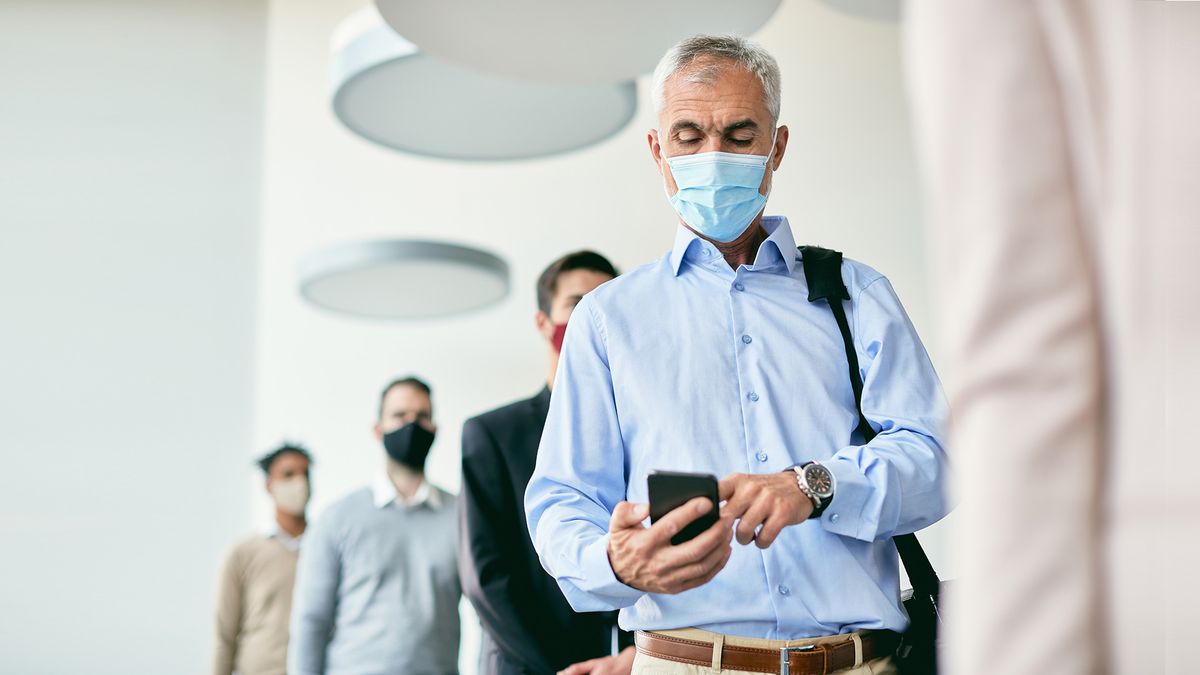 Eine Gruppe von Menschen mit Masken steht in einem Raum in einer Reihe. Der Mann im Vordergrund benutzt ein Smartphone.