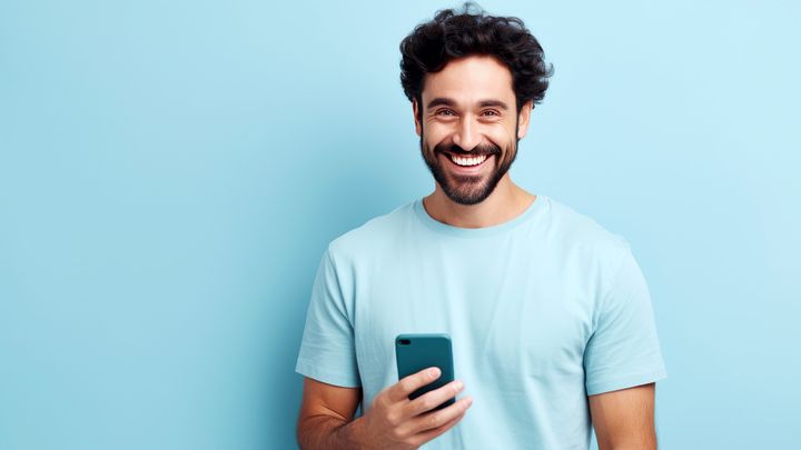 Ein lächelnder Mann mit dunklem Haar und Bart, der ein Smartphone in der Hand hält.