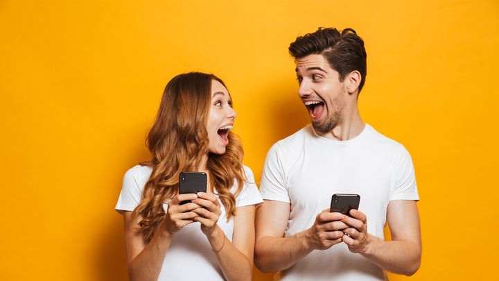 Ein Mann und eine Frau in weißen Hemden blicken sich lachend an, während sie vor einem leuchtend gelben Hintergrund ihre Smartphones in der Hand halten.