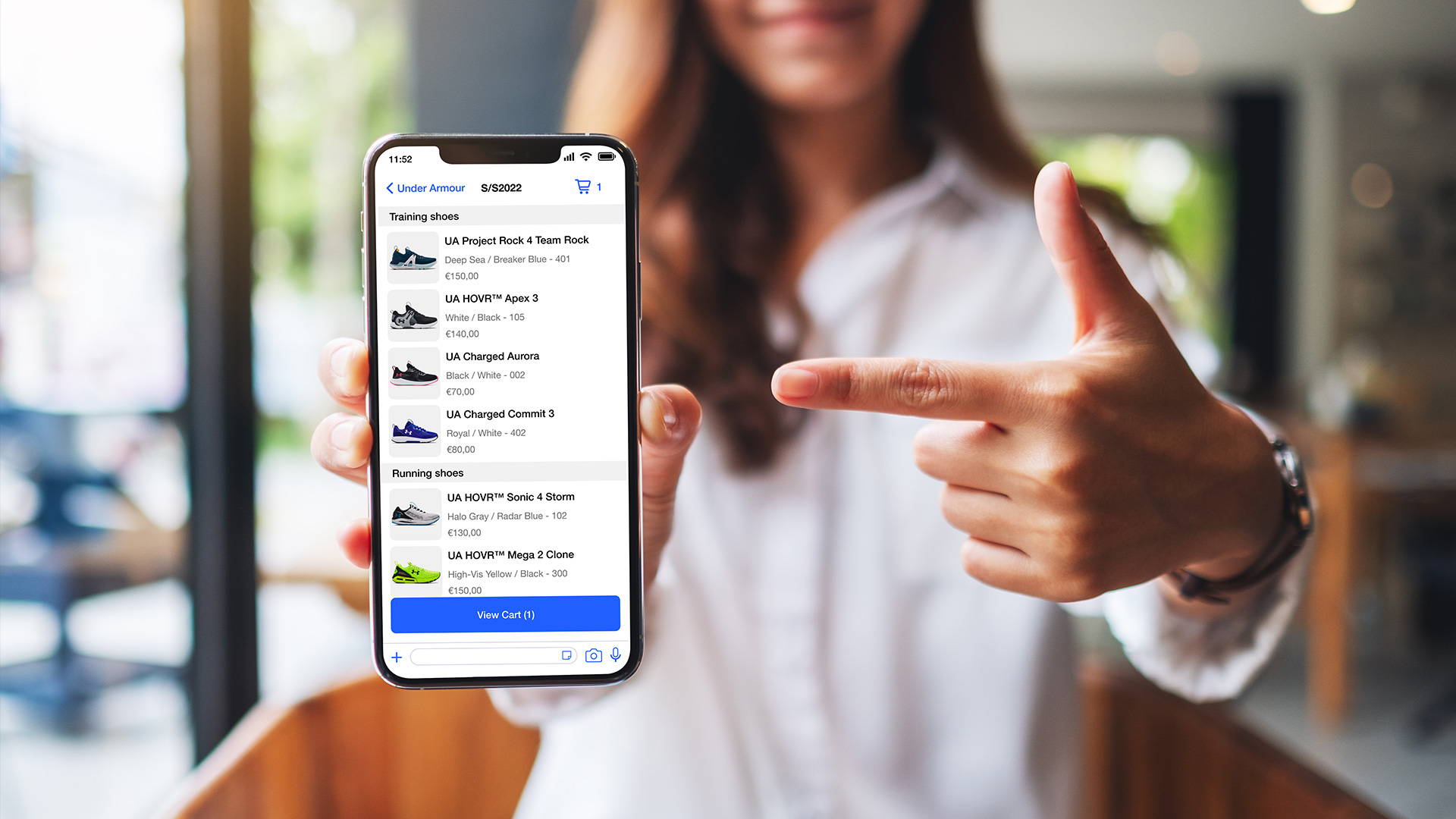 Eine Person zeigt auf den Bildschirm eines Smartphones, auf dem eine Shopping-App mit verschiedenen Trainings- und Laufschuhen angezeigt wird.