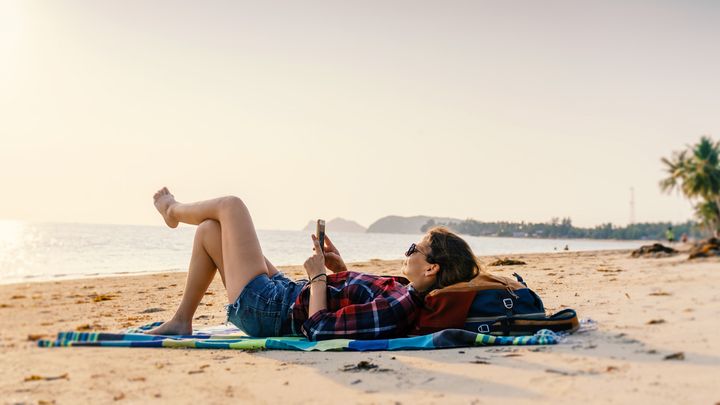 Eine junge Frau liegt am Strand und schaut fröhlich auf ihr Smartphone. Mobile Kanäle wie SMS und WhatsApp bieten viele Up- und Cross-Selling-Möglichkeiten vor Reiseantritt oder direkt am Urlaubsort.