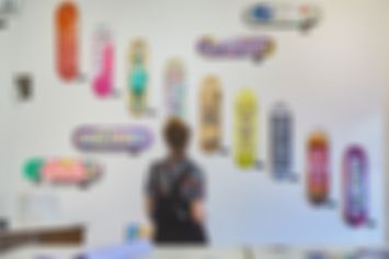 Eine Person steht vor einer weißen Wand und betrachtet die dort hängenden bunten Skateboard Decks.