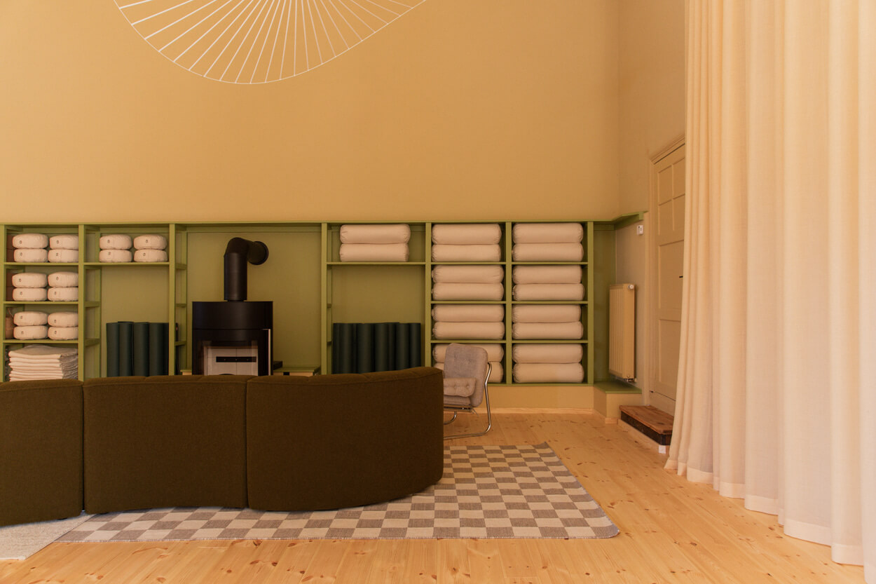 Minimalist room with green shelves holding rolled towels, a black wood stove, curved green sofa, checkered rug, and light wooden floor.