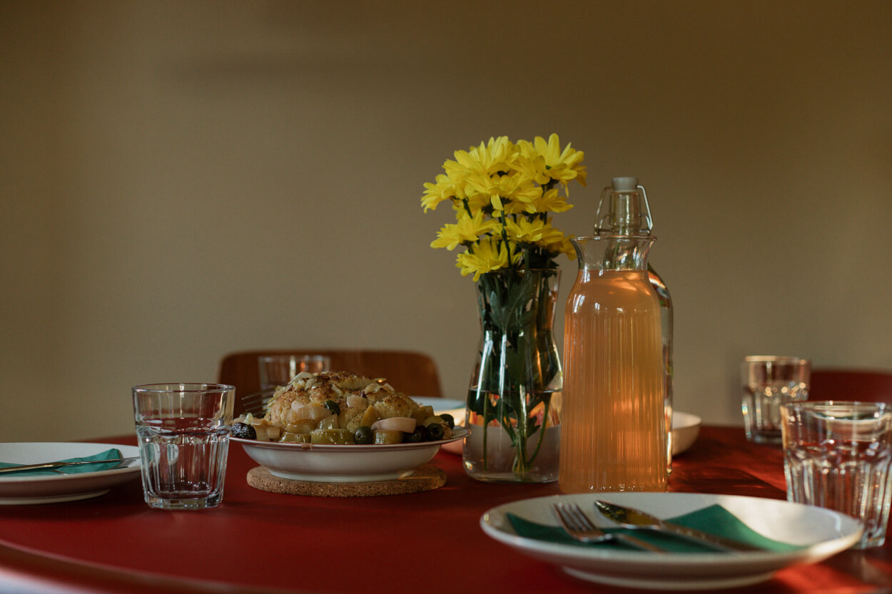 A dining table with a vase of yellow flowers, a glass bottle of juice, plates of food, and glasses on a red tablecloth.
